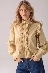 Camisa de color beige con cuello alto, cierre frontal de botones y volados decorativos en el frente, hombros y mangas. Presenta mangas largas con puños ajustados y un ruedo con volado, confeccionada con un diseño romántico y femenino.