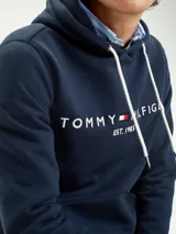 Hoodie azul marino de Tommy Hilfiger con capucha ajustable mediante cordones blancos. Presenta un bolsillo tipo canguro en la parte frontal y el logo de la marca bordado en el pecho junto a la inscripción 'EST. 1985'.