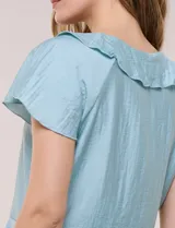 Blusa de satén texturizado color verde menta, con escote en V y volados que bordean el cuello. Presenta mangas cortas acampanadas y doble nivel de volados en la parte inferior, formando una silueta péplum.
