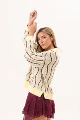 Buzo de punto tipo polo en tono amarillo claro con diseño de ondas a rayas en tonos marrón y celeste. Presenta cuello clásico, cierre frontal parcial con botones y silueta relajada con mangas largas.