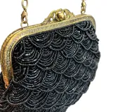 Bolso tipo sobre de estilo vintage, bordado íntegramente con mostacillas negras formando un patrón de ondas. Presenta una boquilla metálica dorada con cierre a presión y una cadena fina dorada para llevar al hombro.