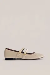 Ballerina flat de mujer confeccionada en tela color beige, con diseño de punta fina y doble tira cruzada con hebillas metálicas doradas. Presenta detalles de ribete marrón oscuro alrededor de la abertura.