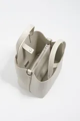 Bolso de mano de tamaño pequeño con estructura rígida, confeccionado en piel reconstituida color gris claro. Cuenta con doble asa superior, correa ajustable con logo de la marca y logo metálico en la parte frontal.