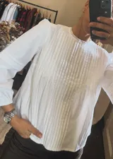 Camisa blanca de algodón con diseño de alforzas verticales en el frente, cuello redondo con detalle de encaje y mangas largas con puños elásticos.