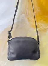 Bandolera Kaira de la línea Ottenta de Carolina de Cunto, color negro con bolsillo frontal dorado texturizado de efecto reptil y arte pintado a mano. Cuenta con correa regulable negra con el logo de la marca en blanco.