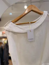 Blusa blanca de diseño cruzado sin mangas, con escote bote y un lazo lateral con hebilla para ajustar en la cintura.