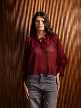 Blusa de plumeti color bordó, con cuello alto ajustable mediante lazo, mangas largas con puños elásticos y detalle de volados en las terminaciones.
