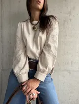 Blusa de color beige con diseño de corte holgado y mangas largas abullonadas. Presenta detalles bordados en tono sobre tono en las mangas y puños ajustados.