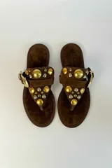 Sandalia plana de gamuza marrón, estilo ojota, con apliques metálicos dorados y hebilla decorativa.