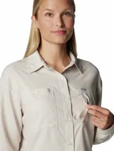 Camisa de senderismo Columbia de manga larga, color beige, con cuello clásico, cierre frontal con botones y bolsillos funcionales en el pecho. Cuenta con ventilaciones traseras de malla para mayor transpirabilidad y tecnología de protección solar y absorción de humedad.