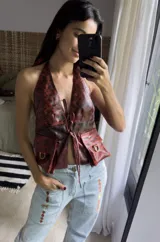 Chaleco tipo top de cuero color óxido con diseño halter, escote profundo y cierre frontal mediante lazos regulables. Cuenta con bolsillos aplicados al frente con detalles metálicos.