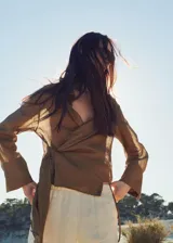 Blusa de ramio color marrón, de diseño recto, con cuello camisero, manga larga con puños abotonados, cierre frontal con botones y detalle de lazo cruzado.