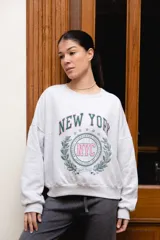 Buzo de felpa gris melange con cuello redondo, mangas largas caídas y estampa frontal estilo universitario con las inscripciones "NEW YORK", "NYC" y "EAST COAST" en tonos verde y rosado.