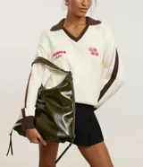 Cartera tipo hobo de gran tamaño confeccionada en poliuretano color negro, con acabado liso y brillante. Presenta un diseño de media luna con cierre superior de cremallera, correa de mano fija y asa larga desmontable. Incluye un detalle de cremallera lateral que permite expandir su capacidad interna.