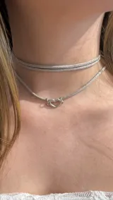 Gargantilla doble estilo choker confeccionada en material tipo gamuza brillante color plateado, con un dije central en forma de corazón calado de metal plateado.
