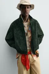Cazadora regular fit de piel con acabado en ante color verde oscuro, de la colección especial Gabriel Azevedo x ZARA. Cuenta con cuello subido, manga larga, bolsillos de vivo en la cadera, bolsillo interior y bajo acabado con elástico. Cierre frontal con cremallera oculta por solapa con botones a presión.