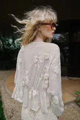 Blusa de escote en pico y manga larga con puños elásticos. Presenta un diseño semitransparente con bordados florales a tono, cintura elástica y cierre frontal mediante lazos con abalorios decorativos.
