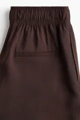 Pantalón culotte marrón chocolate de tiro alto con cintura elástica y lazo para atar. Piernas anchas con bolsillos laterales y un bolsillo decorativo en la parte trasera.