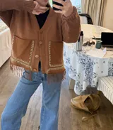 Cardigan de punto color beige con escote en V, cierre frontal con botones, bolsillos delanteros aplicados y detalle de flecos en el ruedo.