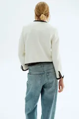 Chaqueta corta de tejido bouclé blanco, con ribetes negros en cuello, frente, bolsillos y puños. Presenta cuello en V, cierre frontal con botones dorados con diseño de nudo y bolsillos insertados.