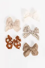Set de cuatro broches para el pelo con diseño de moñas en diferentes texturas y estampados: una moña color crema, una moña a cuadros beige y blanco, una moña marrón con bordado de flores blancas y una moña de lino color marrón.