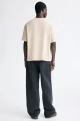 Remera de algodón en color beige con cuello redondo acanalado, hombros caídos y corte holgado. Presenta un estampado frontal con la frase 'RUN WILD' en tonos amarillos y negros con efecto desgastado.