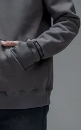 Hoodie oversize color gris oscuro, confeccionado en frisa premium con textura gamuzada. Presenta capucha ajustable con cordones personalizados, bolsillo frontal tipo canguro y puños con abertura para el pulgar.