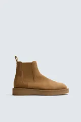 Bota estilo Chelsea de hombre, confeccionada en piel con acabado de serraje en color tostado. Presenta elásticos laterales a tono, horma redonda y suela plana del mismo color.