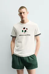 Camiseta de corte relajado confeccionada en piqué de algodón color crudo. Presenta cuello redondo, mangas cortas con ribetes a contraste y estampado frontal con el logo de Italia 90 FIFA World Cup.