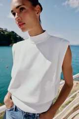 Top de punto color marrón oscuro, con cuello subido, manga sisa con hombreras, pliegues y espalda descubierta con cierre trasero de botones.
