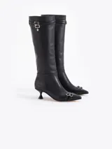 Bota de caña alta confeccionada en cuero vacuno liso, con taco laqueado de altura intermedia y punta fina. Presenta detalles de hebillas metálicas en la caña y en la puntera, incluyendo un dije en forma de corazón.