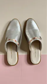 Sandalias chatitas tipo mules, color plateado con detalle de arte en la parte superior y aplique metálico triangular.