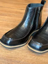 Botas cortas de cuero negro con textura de reptil, diseño de punta cuadrada y suela con detalle de franja en color beige.