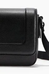 Cartera bandolera negra de forma rectangular, con textura granulada (efecto piel). Presenta una solapa frontal con cierre magnético y una correa ajustable de tela negra.