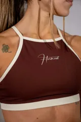 Top deportivo de cuello halter en color marrón chocolate con ribetes en color crema. Presenta el logo de la marca estampado en el centro del pecho.