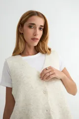 Chaleco de punto con escote en V y cierre frontal de botones. Presenta un diseño de corte recto y textura jaspeada en tono crema.