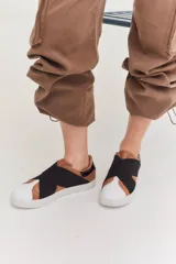 Zapatillas urbanas de color negro con diseño slip-on, caracterizadas por bandas elásticas cruzadas en el empeine y detalles en charol. Cuentan con suela plana de goma al tono.