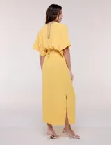 Vestido largo amarillo de corte fluido con escote en V, mangas cortas amplias y apliques de tachas en el escote y puños. Posee cintura elastizada fruncida y falda larga con tajos laterales en el ruedo.
