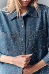 Camisa de denim azul con apliques metálicos plateados y bordado de piedras rojas y negras en los bolsillos delanteros. Tiene calce amplio, cuello camisero y cierre con botones a presión.