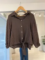 Camisa de lino marrón oscuro con cuello camisero, cierre frontal con botones y mangas largas abullonadas con detalles de bordado broderie en las mangas y en los laterales. Cuenta con un nudo ajustable en la parte inferior delantera.