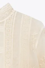 Blusa de manga larga color crema, con cuello alto y detalles de encaje y bordados florales. Presenta paneles verticales con entredoses y un tejido semitransparente.