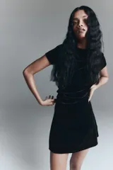 Vestido corto entallado de terciopelo negro, con escote redondo y mangas cortas. Presenta un diseño con pliegues laterales que crean un efecto drapeado en el frente.