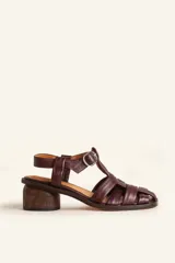 Sandalia de cuero vacuno color marrón oscuro, con diseño de tiras cruzadas estilo romano y taco cilíndrico de madera. Cuenta con tira ajustable con hebilla metálica en el tobillo.