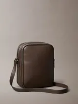 Cartera bandolera Calvin Klein de piel sintética texturizada, color marrón oscuro, con silueta estructurada y correa ajustable. Presenta un bolsillo exterior frontal y un compartimento principal con cierre de cremallera.