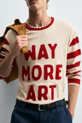 Camiseta de manga larga con cuello redondo, cuerpo color beige claro y mangas a rayas horizontales rojas y beige. Presenta texto frontal en relieve color rojo que dice "WAY MORE ART".