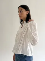 Blusa de brodery color blanco, con cuello redondo, mangas largas abullonadas con puños elásticos y cierre frontal con botones forrados. Presenta detalles de puntillas verticales en el frente.