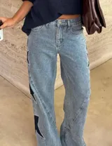 Jean de tiro alto con pierna amplia de corte recto, confeccionado en denim de lavado claro. Presenta aplicaciones de estrellas en contraste con bordes decorados con tachas metálicas.