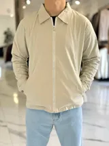 Campera de gabardina beige de corte boxy fit, con cuello camisero, cierre frontal de cremallera blanca y bolsillos laterales ribeteados.