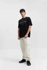 Remera de manga corta color beige, con escote redondo y fit regular. Presenta un estampado frontal en color marrón con la palabra "Resilience" en tipografía gótica y el texto "Original The World Has Changed Established Since 1976 // NLD Mistral".