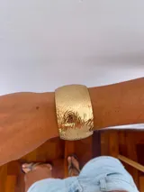 Brazalete tipo cuff de metal dorado con diseño de relieve ondulado y acabado brillante. Es de diseño abierto y ajustable.